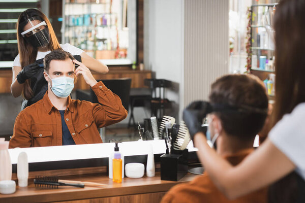 mirror reflection of hairdresser in face shield cutting hair of man pointing with fingers, blurred foreground