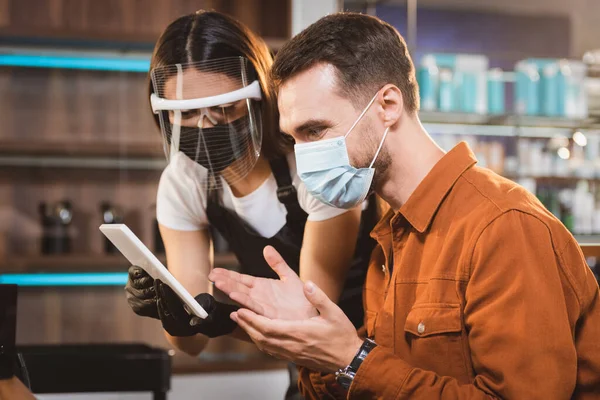 hairdresser in face shield holding digital tablet near client in medical mask pointing with hands