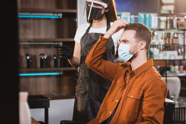 man in medical mask touching his hair near barber in protective equipment pointing with finger