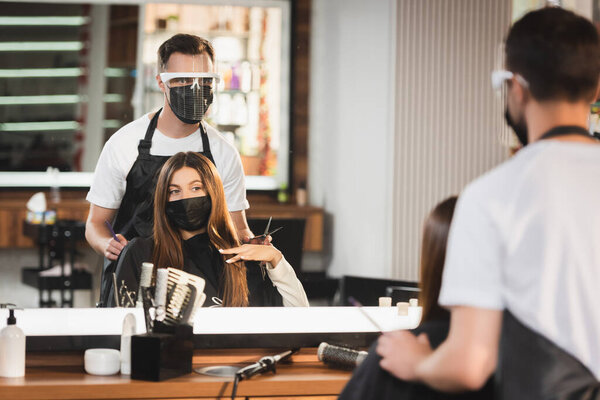 mirror reflection of barber in protective equipment near client in medical mask, blurred foreground
