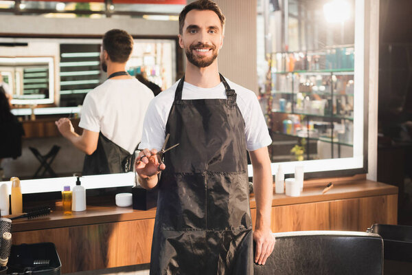 smiling hairdresser in apron holding scissors while standing at workplace on blurred background
