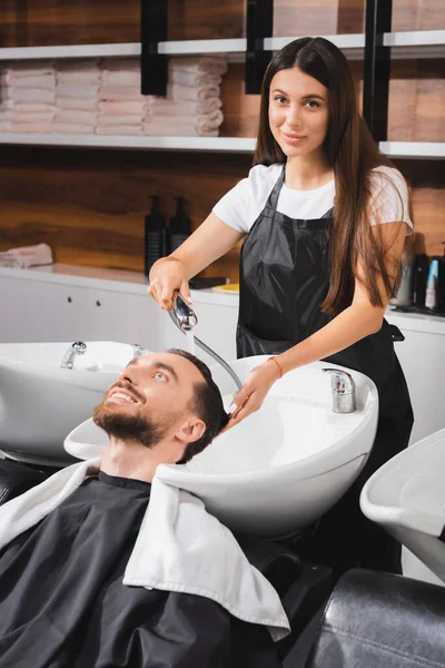 Jovem Barbeiro Avental Olhando Para Câmera Enquanto Lava Cabelo Cliente — Fotografia de Stock