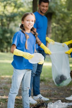 girl holding plastic plate and looking at camera near father with recycled bag on blurred background, ecology concept clipart