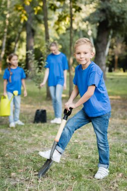boy digging ground with shovel near mother and sister with watering can and young seedling on blurred background, ecology concept clipart