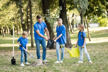 Çocuk toprağı kazıyor, kız su kovası tutuyor ve ebeveynler genç ağaç taşıyor, ekoloji konsepti.