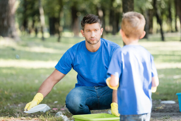 человек собирает пластиковый мусор в парке рядом с сыном на размытом переднем плане, экологическая концепция