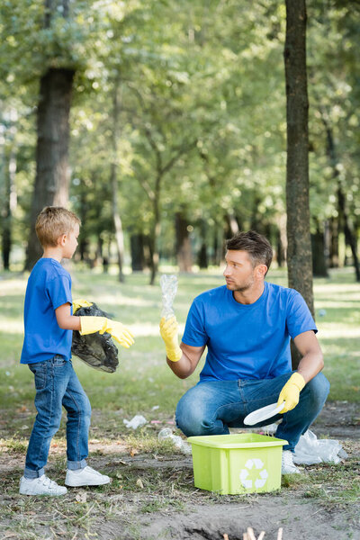 father and son collecting plastic garbage into container with recycling emblem, ecology concept