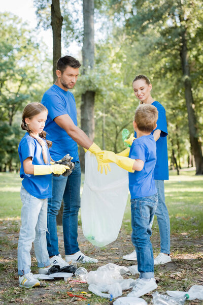 Семья волонтеров в резиновых перчатках собирает мусор в переработанном пластиковом пакете в лесу, экологическая концепция