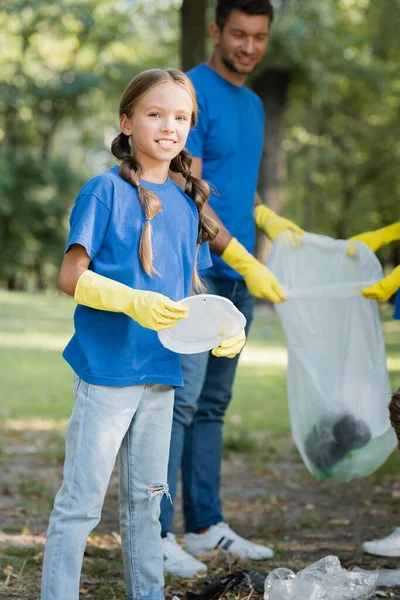 Girl Holding Plastic Plate Looking Camera Father Recycled Bag Blurred — Stock Photo, Image