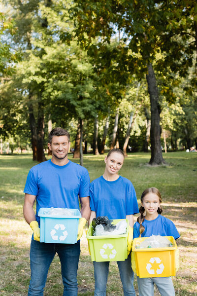 smiling volunteers family holding containers with recycling emblems, full of plastic rubbish, ecology concept