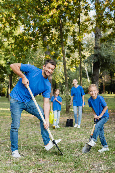 father and son smiling at camera while digging ground, and mother with daughter holding young tree and watering can on blurred background, ecology concept