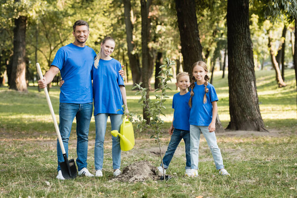 счастливая семья волонтеров, стоящих возле посаженного молодого дерева с лопатой и лейкой, экологическая концепция