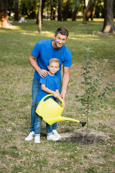 boy looking at camera while watering planted young tree near smiling father, ecology concept