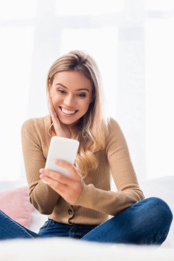 Yatak odasındaki bulanık ön planda akıllı telefon kullanan mutlu kadın.  