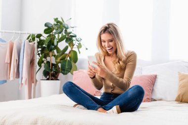 Yatakta bacak bacak üstüne atmış otururken akıllı telefon kullanan heyecanlı bir kadın. 