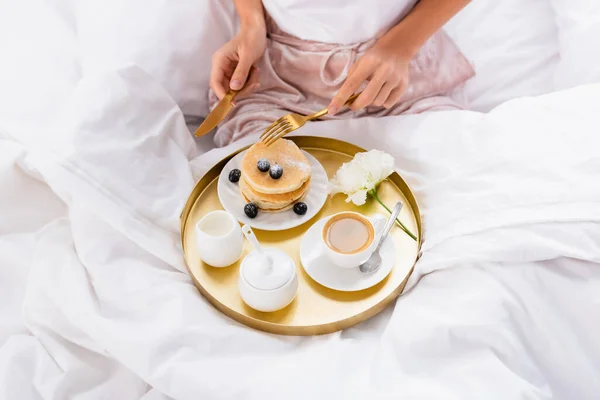 ベッドの上で朝食トレイにパンケーキ コーヒー カーネーションの近くに刃物を保持する女性の作物ビュー — ストック写真