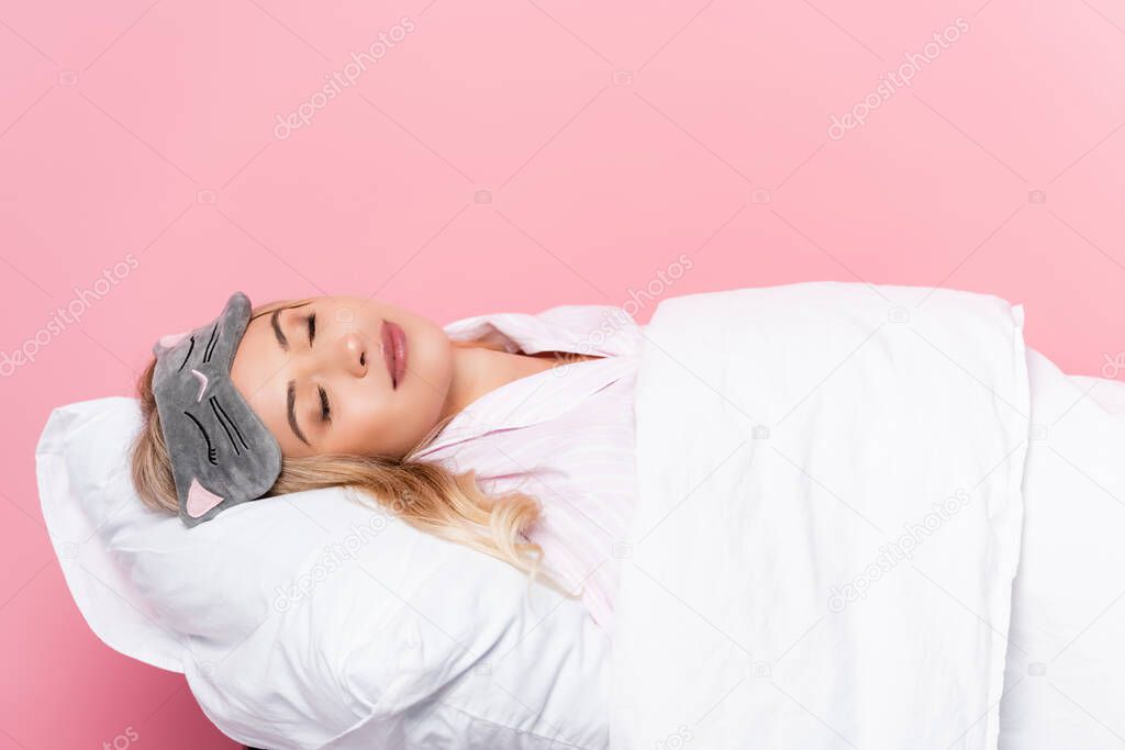 Young woman in blindfold sleeping on pillow with blanket on pink background