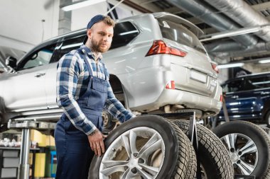 young mechanic looking at camera near wheel and auto raised on car lift clipart