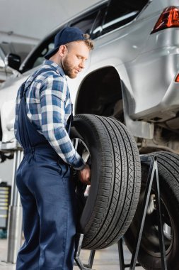 mechanic in overalls holding wheel near automobile raised on car lift on blurred background clipart
