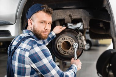 young mechanic looking at camera while adjusting wheel hub on blurred background clipart