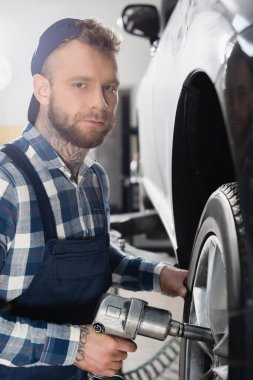 young mechanic looking at camera while fixing car wheel with pneumatic wrench clipart