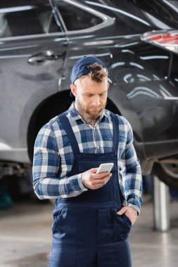 mechanic in overalls holding hand in pocket while chatting on smartphone on blurred background clipart