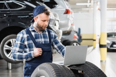 young technician working on laptop near cars in workshop on blurred background clipart