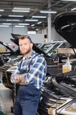 repairman in uniform standing with crossed arms and looking at camera near cars with opened hoods clipart