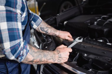 Ön planda araba motoru kompartımanının yanında İngiliz anahtarı tutan dövmeli teknisyenin kısmi görüntüsü