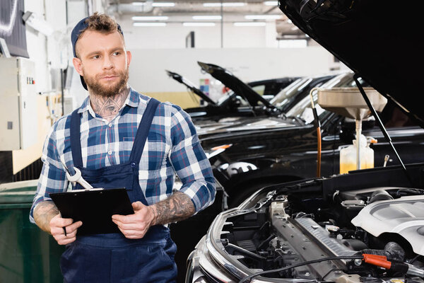 young repairman looking away while standing with clipboard near car with opened hood