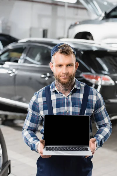 mechanic holding laptop with blank screen near automobiles on blurred background