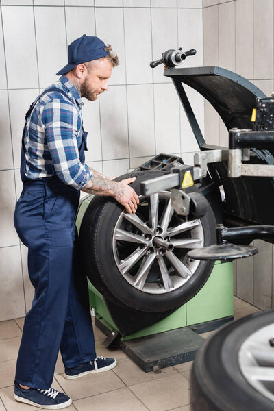 young tattooed mechanic checking wheel on balance control machine in workshop on blurred foreground