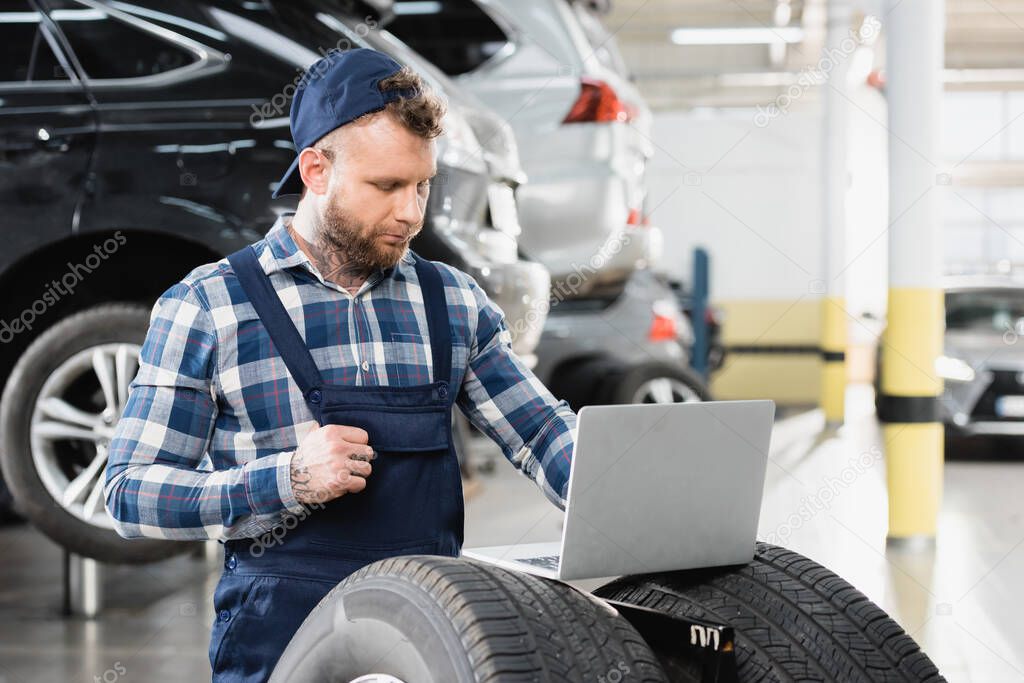 giovane tecnico che lavora su laptop vicino alle auto in officina su sfondo sfocato