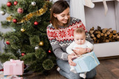 smiling woman holding present near infant son and decorated christmas tree
