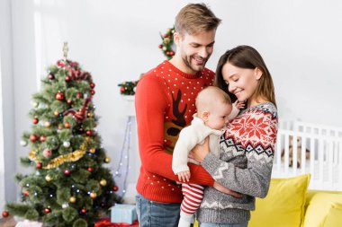 Mutlu ebeveynler kollarını tutuyor bebeğim. Arka planda süslenmiş Noel ağacının yanında. 
