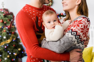 Anne ve babasının kollarında yeni doğmuş bir çocuk ve arka planda bulanık Noel ağacı.