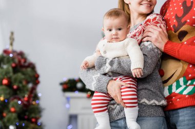 Kocasının yanında kucağında mutlu bir kadın ve arka planda bulanık bir Noel ağacı var.