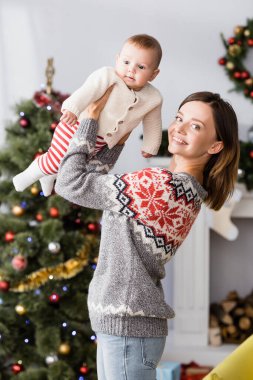 Arka planda bulanık noel ağacının yanında kollarını tutan mutlu bir kadın.