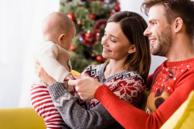 Neşeli ebeveynler arka planda bulanık Noel ağacının yakınındaki bebeğe bakıyorlar.