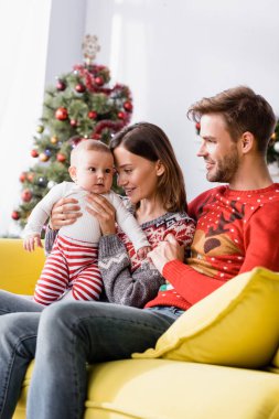 Kollarında mutlu bir kadın, süveter giyen neşeli bir koca ve arka planda bulanık bir Noel ağacı olan bir çocuğun yanında.
