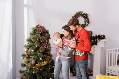 Mutlu bir kadın kollarını tutuyor. Neşeli bir koca ve süslü bir Noel ağacının yanında.