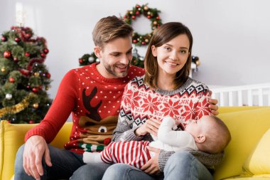Süveter giyen mutlu ebeveynler bulanık arka planda noel ağacıyla küçük oğlunun yanında gülümsüyor. 