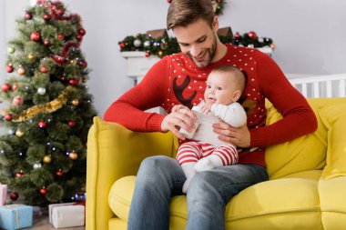 Mutlu bir baba bebeğini beslerken elinde anne sütüyle arka planda bulanık bir Noel ağacı tutuyor. 