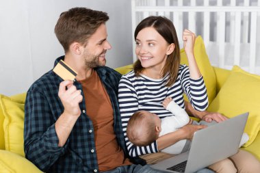 cheerful man holding credit card near excited wife with infant son while online shopping  clipart