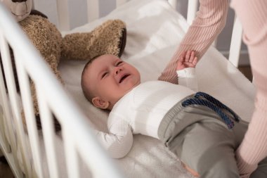 Anne, ön planda bebek beşiğinde ağlayan bebeği kaldırıyor. 