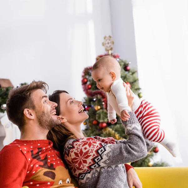 背景にぼやけたクリスマスツリーの近くにいる赤ちゃんの息子を見ている明るい両親の側の見解 — ストック写真