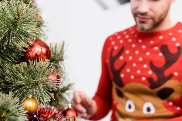 cropped view of bearded man in sweater decorating christmas tree on blurred foreground