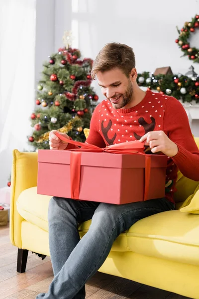 Happy Man Red Sweater Looking Wrapped Present While Sitting Sofa — Stock Photo, Image