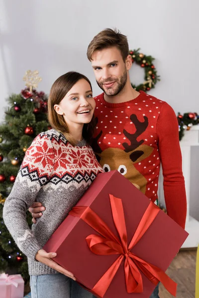 Pleased Man Red Sweater Hugging Excited Wife Holding Present Christmas — Stock Photo, Image