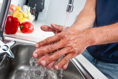 Mutfakta, arka planda sebzelerin yanında ellerini yıkayan adam görüntüsü bulanık. 